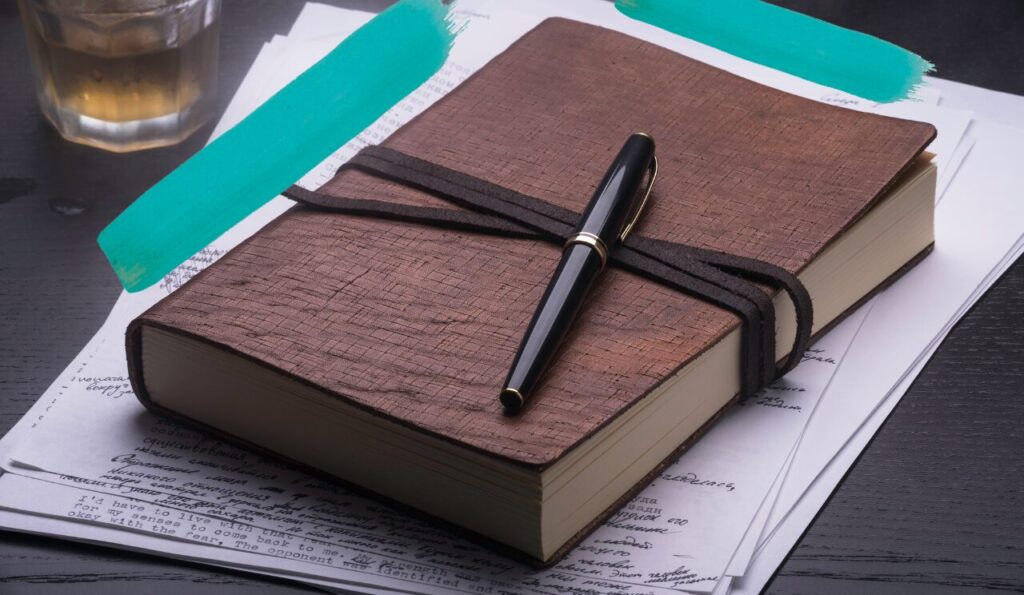 example of a man's career journal. A leather bound, weighty looking journal with a pen lay on top of it. In the background, a glass of whiskey is visible.